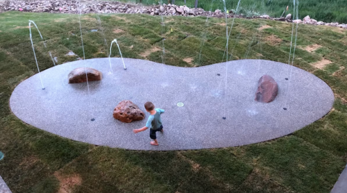 A child plays on a stone-patterned splash pad with water jets, surrounded by grass and rocks.